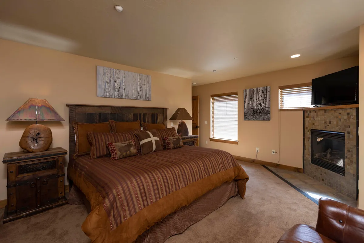 A spacious bedroom with a large bed adorned in striped bedding, a wooden headboard, and a glowing fireplace. Decorative artwork adds warmth, and the scene feels restful and inviting.
