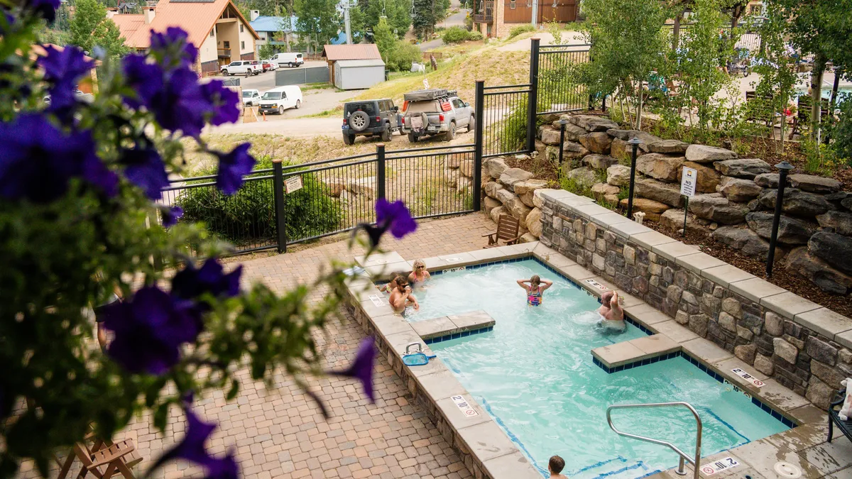 This exterior image presents a vibrant swimming pool scene with people relaxing in the water, surrounded by colorful flowers and lush natural landscaping. The backdrop features mountain scenery that hints at the property's resort setting, and the overall atmosphere suggests active enjoyment and relaxation just steps from the home.