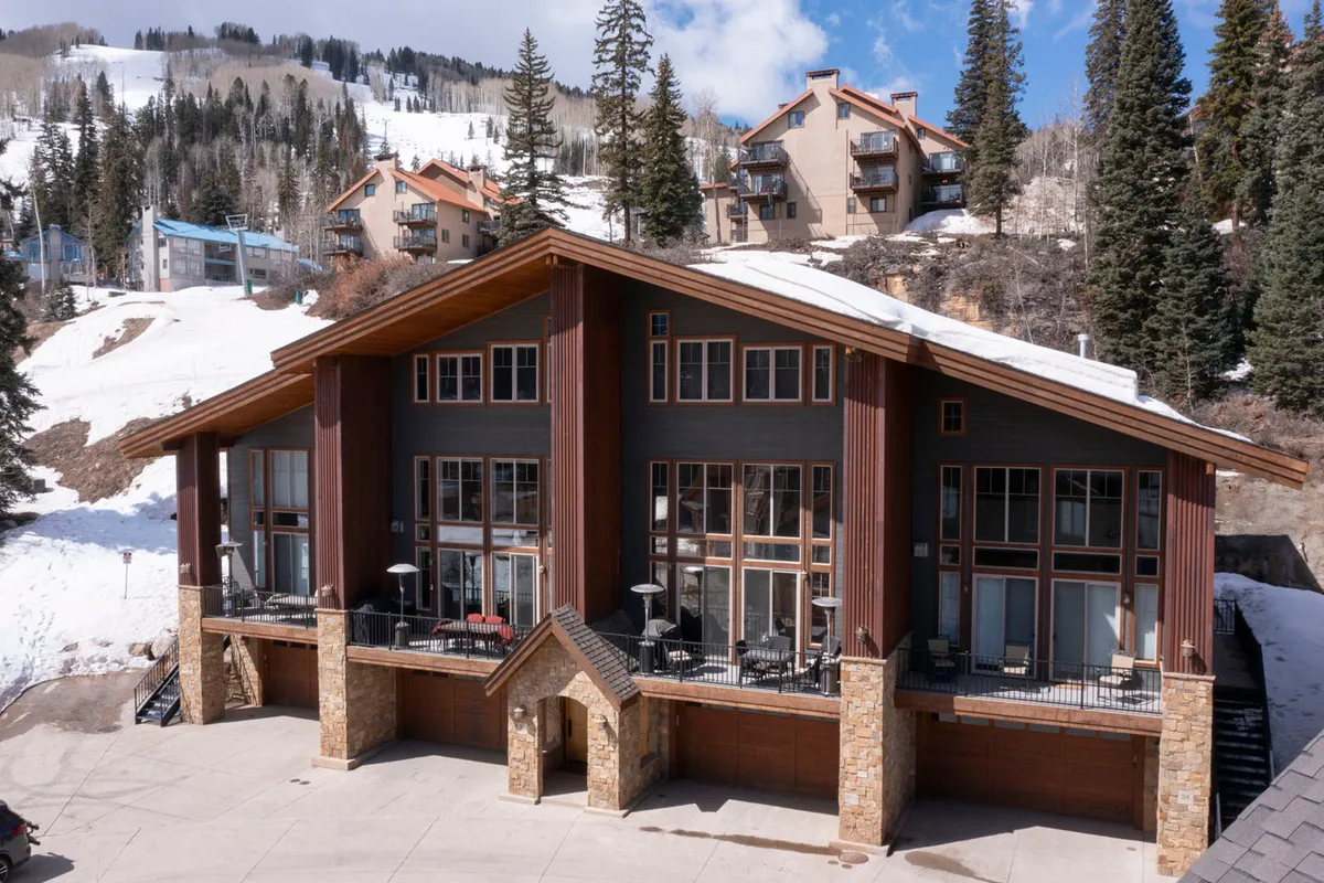 An exterior scene showing the lodge's large windows, stone facade, wooden accents, and a snow-covered landscape. The patio area, surrounded by snow, offers a sense of proximity to the slopes and mountain activity.