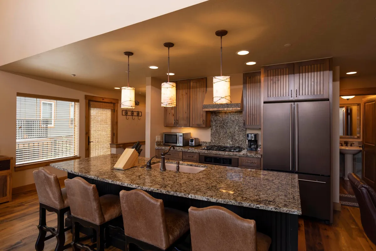 A close-up view of the kitchen area showcasing a sleek granite countertop, pendant lights, stainless steel appliances, and warm wooden cabinetry. The layout is modern and functional, ideal for prepping meals with ample workspace and contemporary amenities.