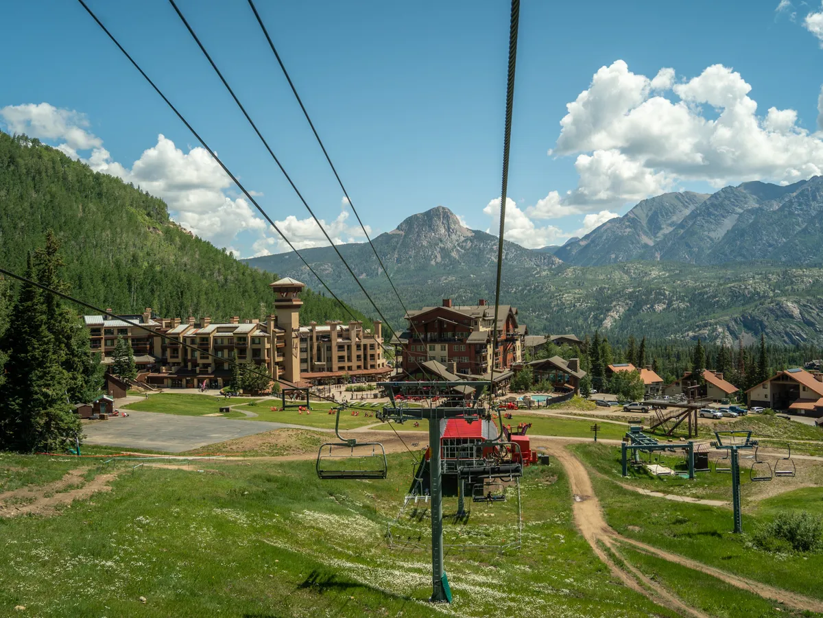 A panoramic exterior view of ski resort buildings set against a dramatic mountain backdrop. A ski lift is visible, hinting at easy access to the slopes, while lush green grass in the foreground suggests year-round activities. The image captures the excitement and convenience of true ski-in/ski-out access.
