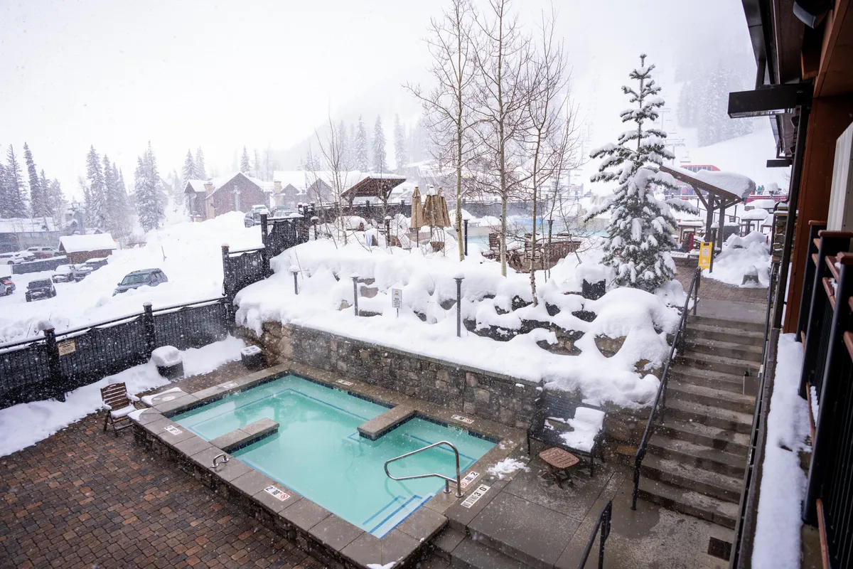 An inviting outdoor hot tub sits amidst a snow-covered landscape, surrounded by tall evergreen trees and flanked by snowy stairs. The setting exudes a sense of winter adventure and group relaxation, with the hot tub providing a prime spot to enjoy the surrounding mountain scenery and socialize after a day on the slopes.