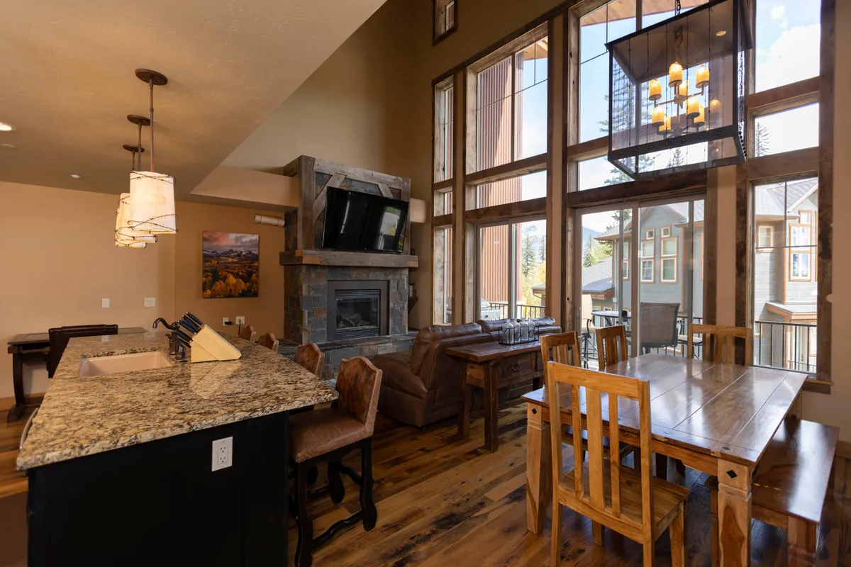 A welcoming open-concept space with large windows letting in natural light, a stone fireplace, a dining table adjacent to the kitchen island, and comfortable furnishings. The setting encourages both family gatherings and quiet, cozy meals, with a versatile layout that keeps everyone connected.