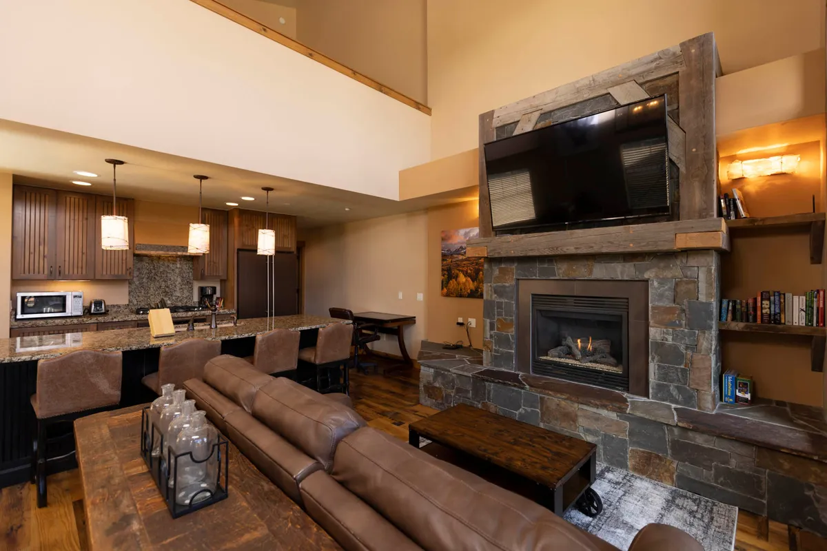 A cozy common area with a fireplace, a comfortable sofa, kitchen island, television, and a visible bookshelf. The space is arranged for relaxation, group gatherings, movie nights, or reading by the fire.