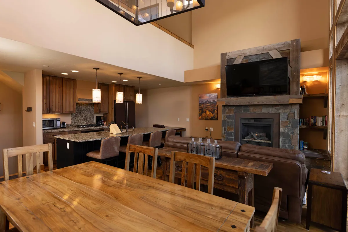 A spacious kitchen and dining area featuring a large wooden dining table that comfortably seats a group, a stone fireplace adding a cozy ambiance, a kitchen island with bar stools, and modern overhead light fixtures. The scene is set for family dinners, social gatherings, and shared meals, with the kitchen seamlessly connected to the dining space.