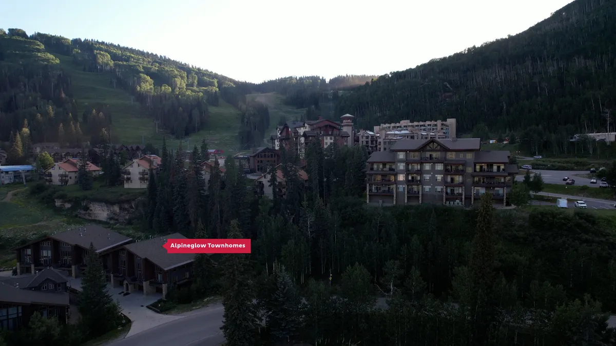 This image displays the Alpineglow Townhomes nestled within a scenic mountain landscape, with other resort buildings nearby and the expansive mountains forming a breathtaking backdrop. The scene evokes a sense of being right at the heart of family-friendly adventure, with easy access to both the slopes and nature trails.