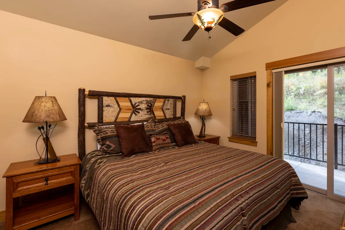 A warmly lit bedroom featuring a bed with decorative pillows and a rustic headboard. Nightstands with lamps sit on either side, and a sliding glass door opens to an outdoor view, creating a tranquil, nature-infused retreat.