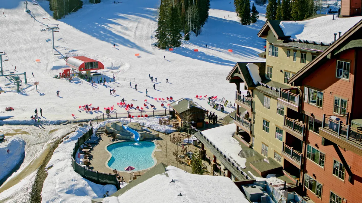 A bustling exterior view of the ski slopes with skiers, a ski lift, a swimming pool, and the lodge in the background. The image captures the excitement and activity just outside the property, with clear sightlines to the ski action.