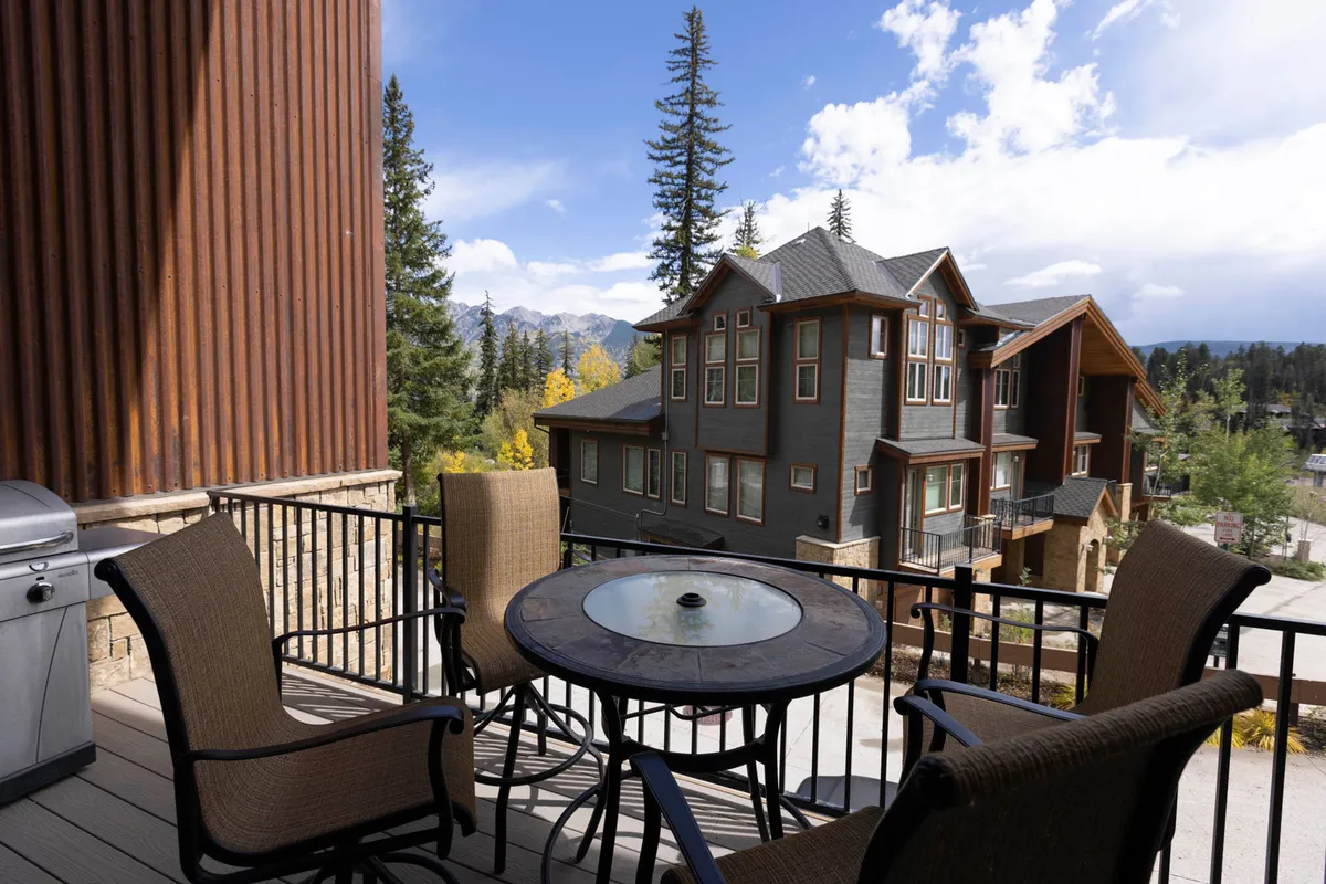 An exterior balcony featuring comfortable furniture overlooks a picturesque mountain view, with a neighboring house and lush natural surroundings in sight. The scene invites visions of après-ski hot cocoa or summer evening barbecues with family.
