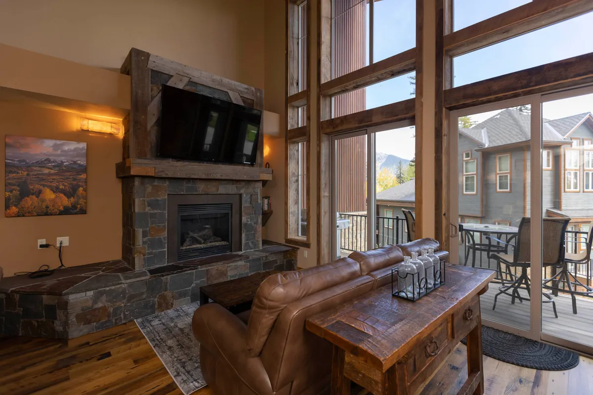 An inviting living area with a leather sofa, a glowing fireplace, and expansive windows that flood the space with natural light. The windows offer a glimpse of the outdoors, reinforcing the connection between the cozy interior and the mountain landscape just outside.
