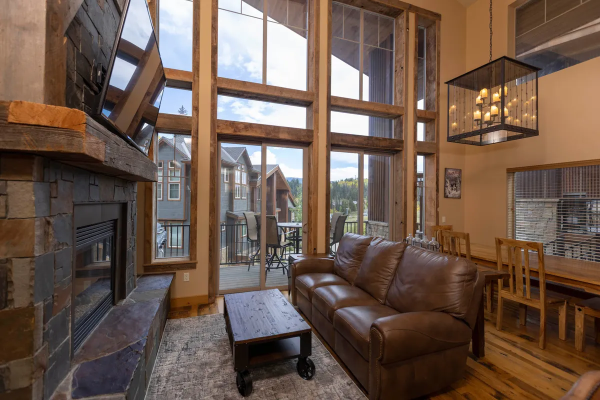 A spacious living area featuring large windows with sweeping scenic views, a comfortable leather sofa, a stone fireplace, and an adjoining dining area with a wooden table. Natural light fills the space, and the fireplace is clearly the focal point for cozy gatherings.